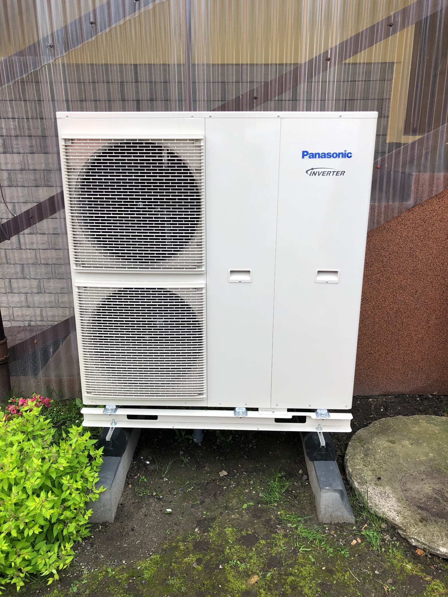 Biała zewnętrzna jednostka klimatyzacyjna Panasonic Inverter, zamontowana na betonowych podstawach, widoczna na tle ściany z blachy falistej i brązowego tynku.
