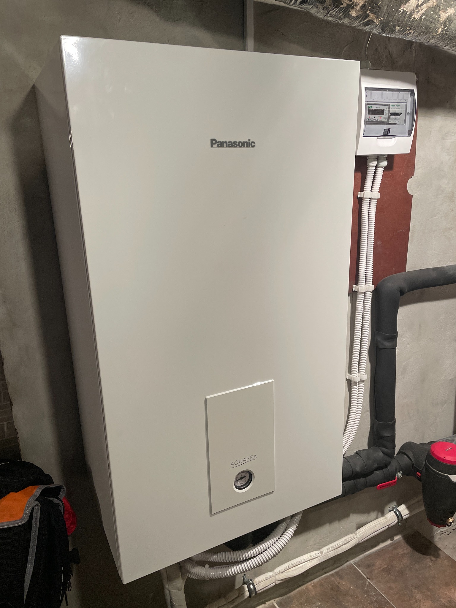 Biała jednostka wewnętrzna z logo Panasonic Aquarea, zamontowana na szarej ścianie w kotłowni, widoczne rury instalacyjne i skrzynka elektryczna.