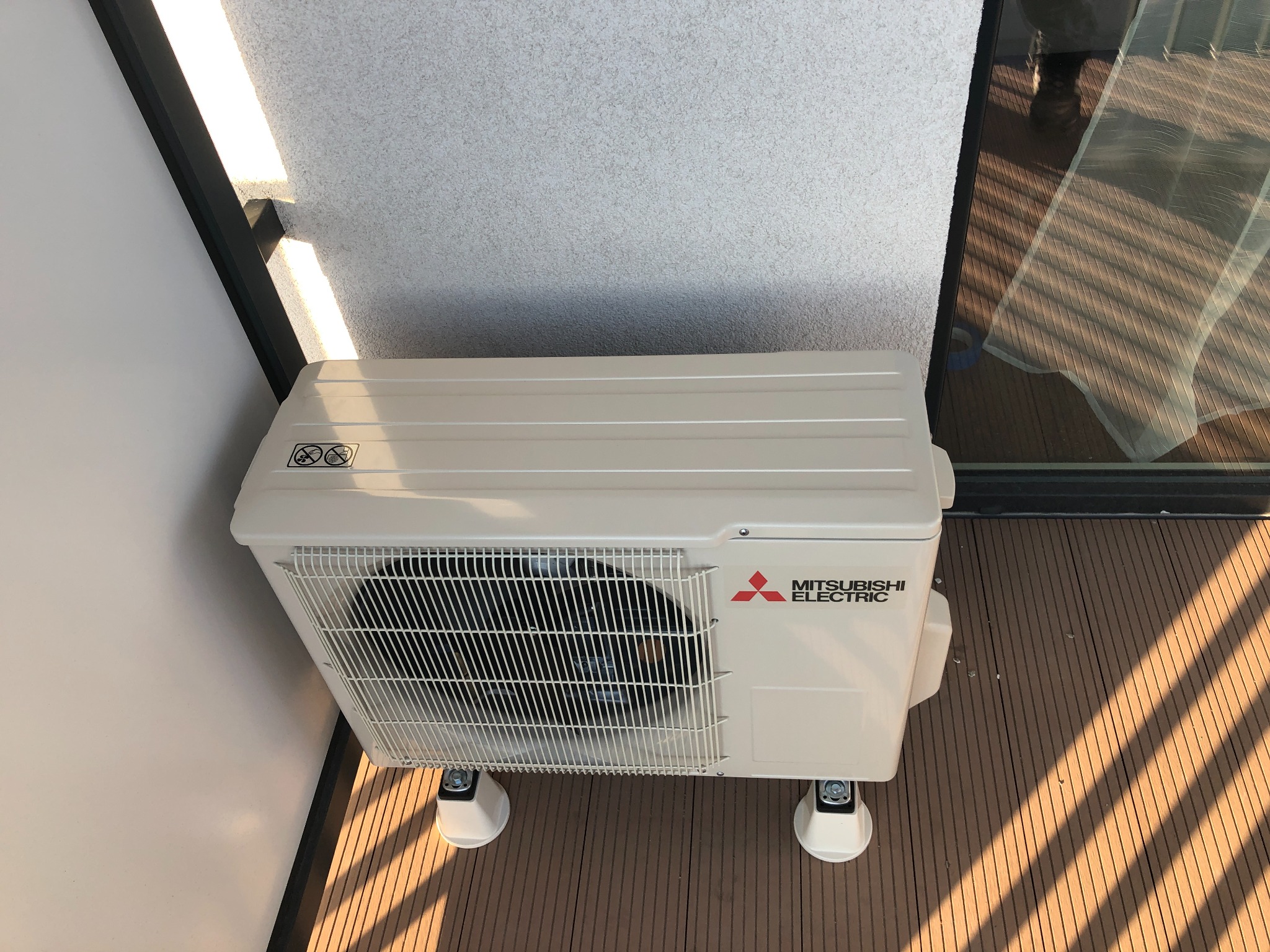 Zewnętrzna jednostka klimatyzacji Mitsubishi Electric zamontowana na balkonie z drewnianą podłogą, widoczna z góry pod kątem, z fragmentem okna i ściany w tle.