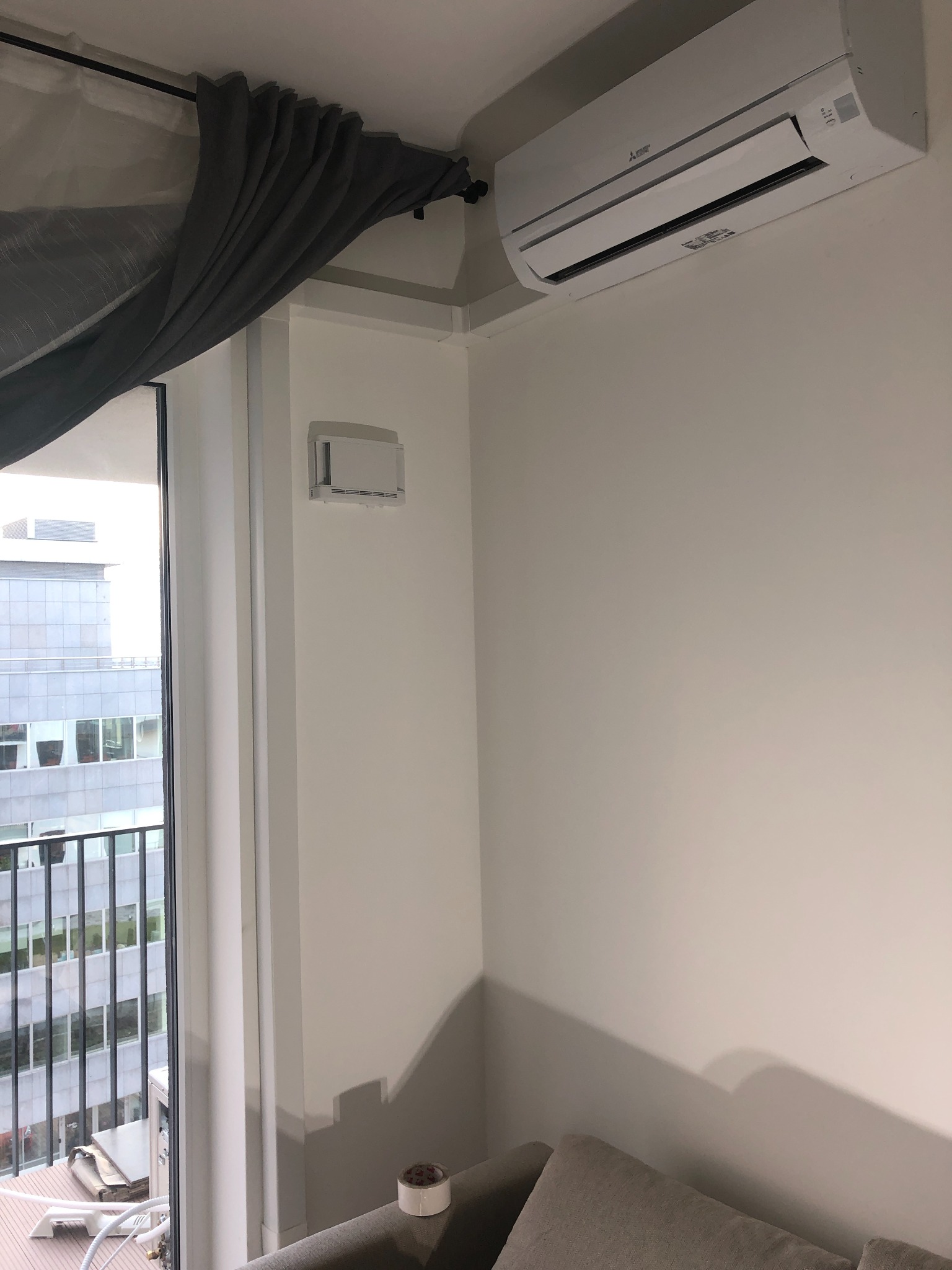Wnętrze pokoju z zamontowaną jednostką klimatyzacji Mitsubishi na ścianie, widoczny również nawiewnik, szare zasłony i fragment sofy w dolnym rogu, za oknem budynek biurowy.