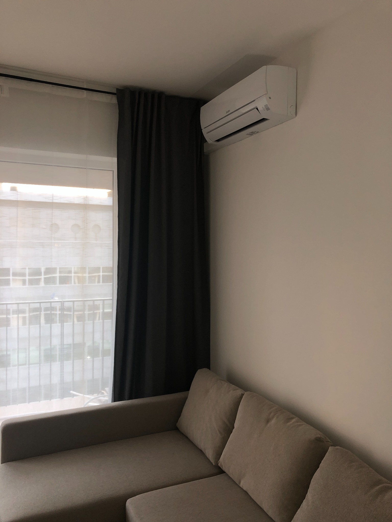 Biały klimatyzator ścienny zamontowany nad szarą sofą w nowoczesnym salonie, z widokiem na budynek przez okno z zasłoną.