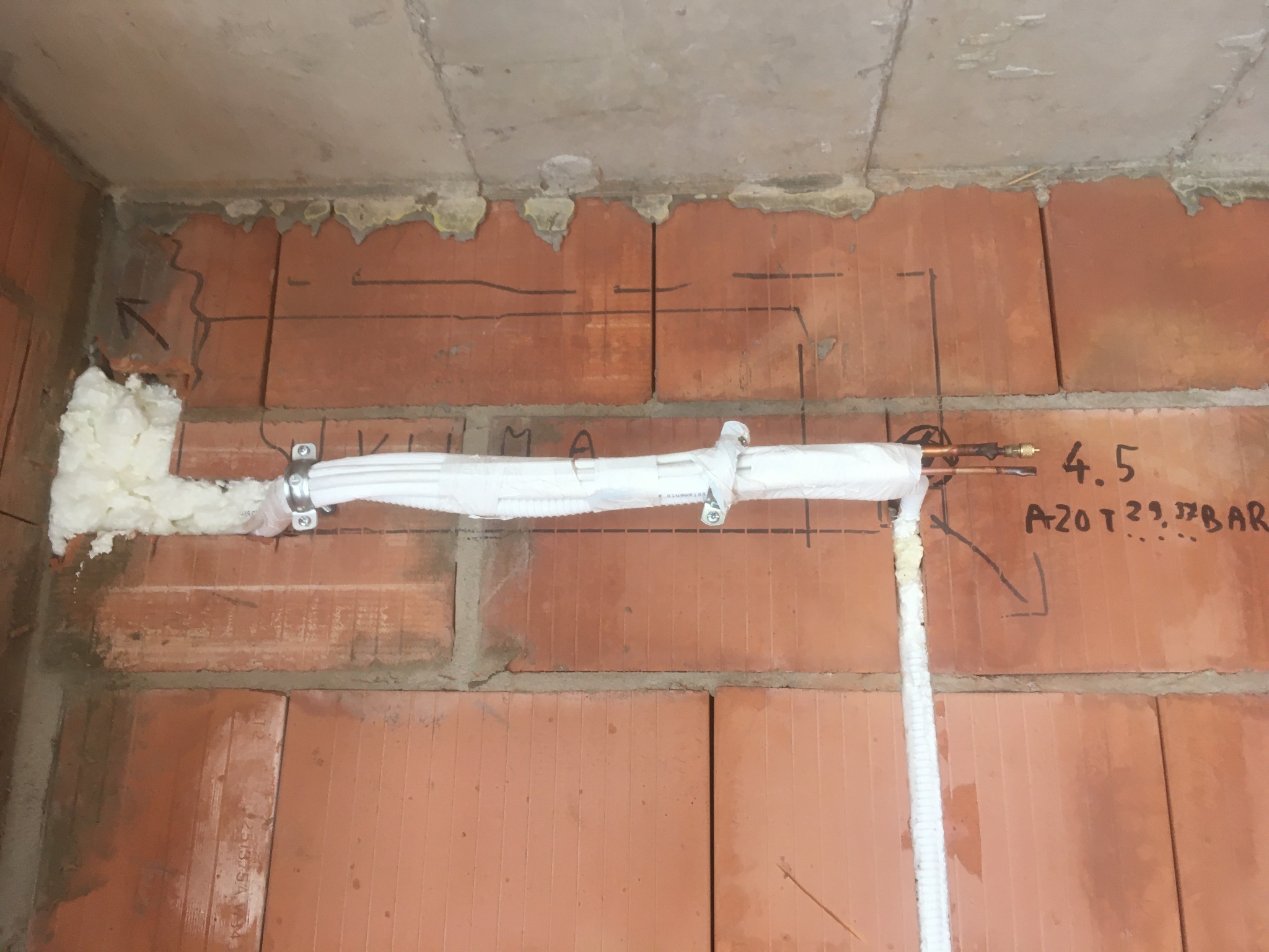 Rury w białej izolacji zamocowane do ściany z czerwonej cegły z widocznymi odręcznymi notatkami dotyczącymi parametrów, pianka montażowa w szczelinach, widok instalacji w trakcie prac.