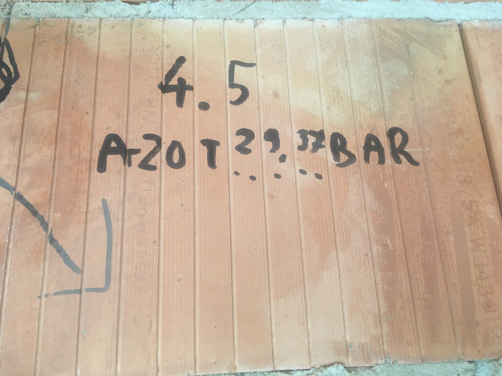 Fragment muru z cegieł ceramicznych, na którym czarnym markerem naniesiono odręczne notatki techniczne dotyczące ciśnienia i oznaczenia gazu: 4.5, A-20 T 29,37 BAR, strzałka.