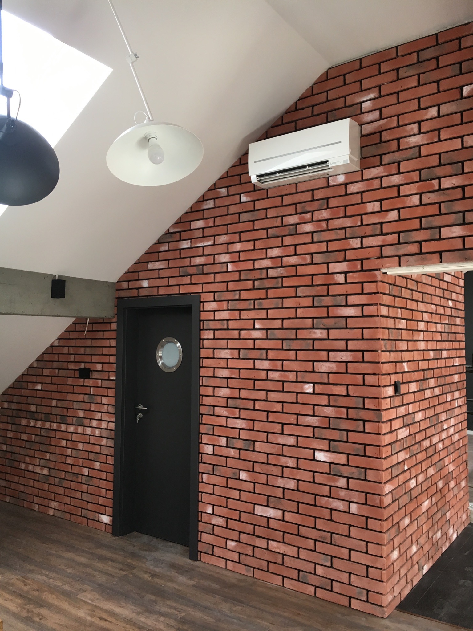Wnętrze poddasza z czerwoną cegłą na ścianach, gdzie zamontowano biały klimatyzator ścienny obok czarnych drzwi z okrągłym okienkiem.
