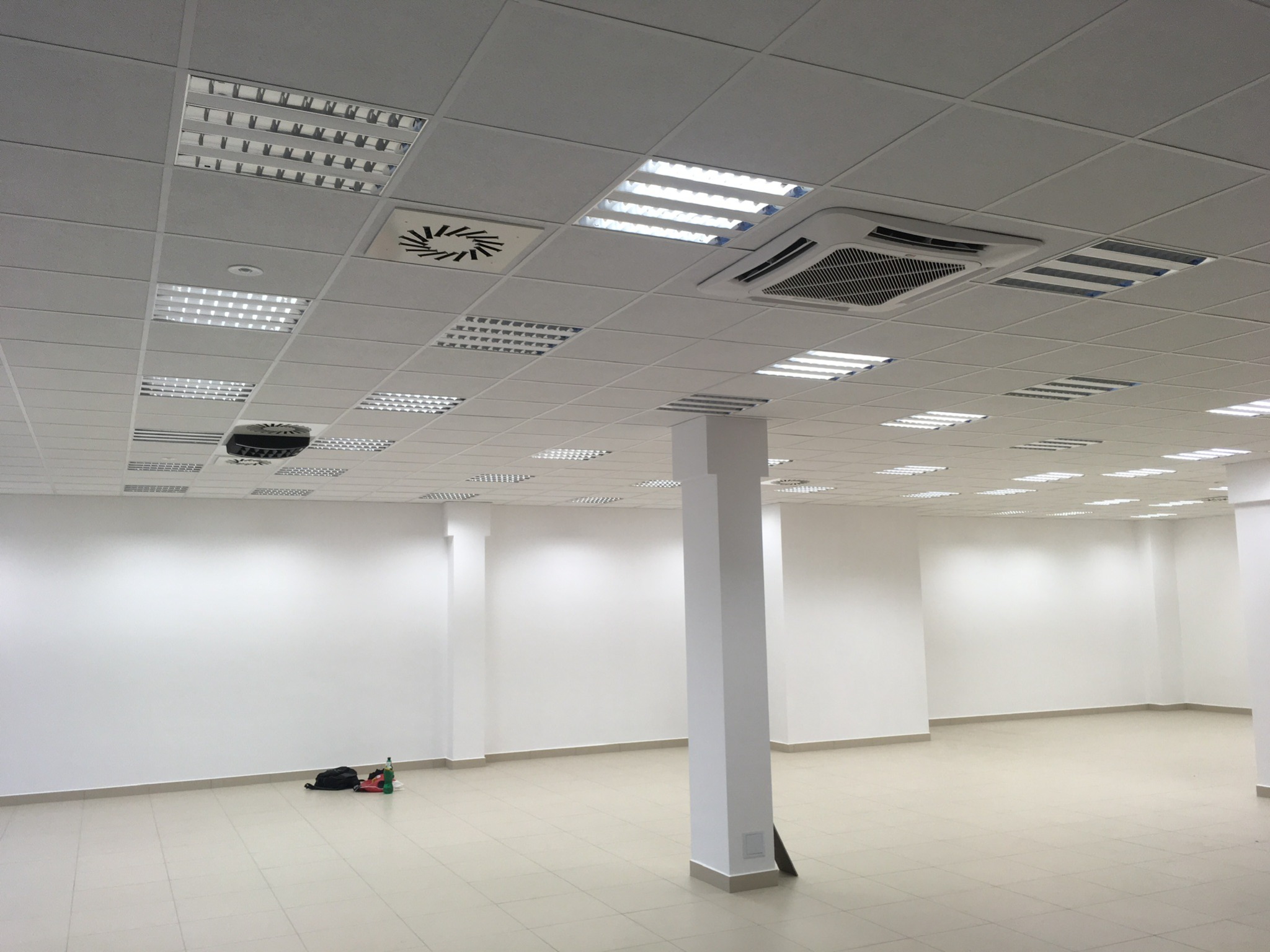 Puste biuro z białymi ścianami i sufitem kasetonowym, widoczne lampy, kratki wentylacyjne oraz klimatyzator kasetonowy. Na podłodze leżą torba i butelka.