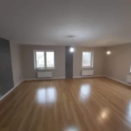 Puste, jasne pomieszczenie po remoncie z panelami podłogowymi, szarą ścianą akcentującą i trzema oknami z grzejnikami pod parapetami. Widoczne niedokończone gniazdka elektryczne na ścianie.