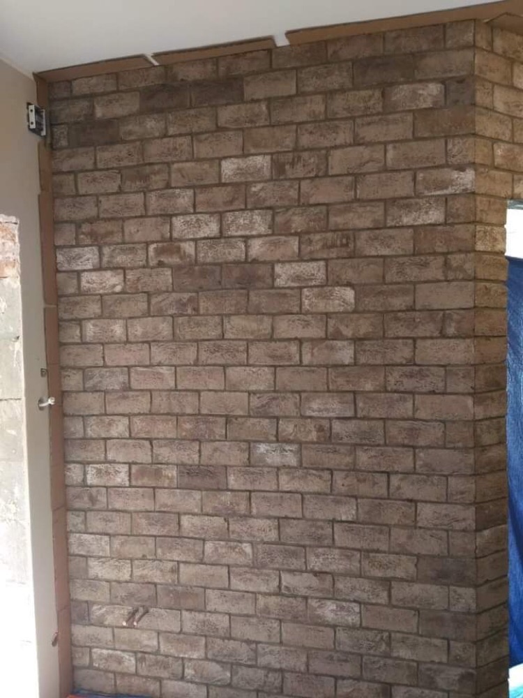 Ściana wykończona cegłą dekoracyjną w odcieniach brązu i szarości, widoczne elementy montażowe przy suficie i drzwiach.