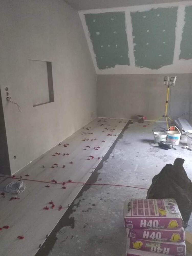 Poddasze w trakcie remontu: położona częściowo podłoga z płytek z czerwonymi krzyżykami dystansowymi, worki z klejem H40, ściany przygotowane do malowania, gipsowane płyty na skosach dachu, lampa...