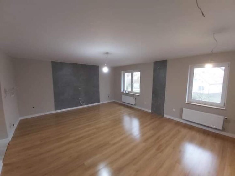 Wykończone wnętrze pokoju z panelami podłogowymi i świeżo pomalowanymi ścianami w odcieniach beżu i szarości, widoczne dwa okna z grzejnikami oraz wiszący przewód od lampy.