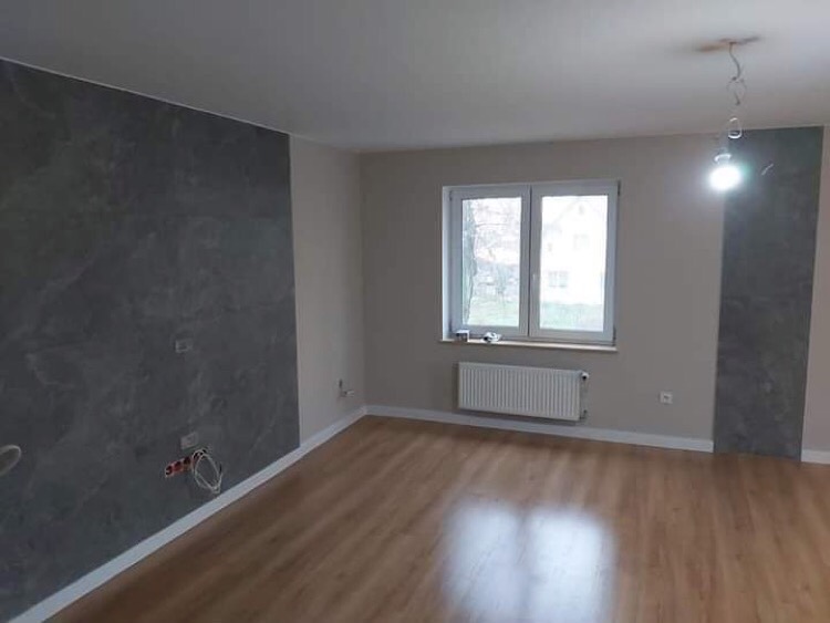 Pusty pokój po remoncie z panelami imitującymi kamień na ścianie, widoczne niedokończone podłączenia elektryczne, drewniana podłoga i grzejnik pod oknem.