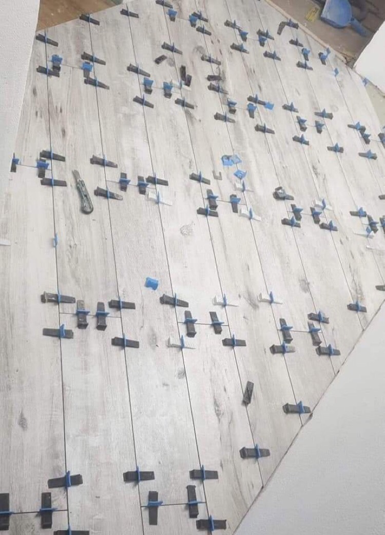 Układanie jasnoszarych płytek podłogowych imitujących drewno, z użyciem systemu poziomowania z niebieskimi klipsami i sznurkami, widoczny nóż do docinania płytek.