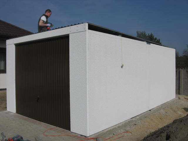 Montaż blaszanej konstrukcji garażu z brązową bramą segmentową; pracownik na dachu przykręca elementy, widoczne narzędzia i kabel zasilający na ziemi.