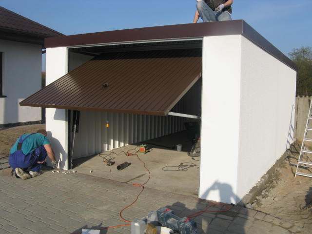 Montaż garażu blaszanego z bramą segmentową w kolorze brązowym, widoczny pracownik uszczelniający szczelinę przy posadzce, drugi pracownik siedzi na dachu.