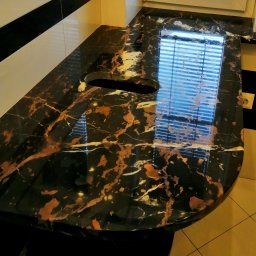 VIPoler Marcin Wiśniewski - Czarny blat kamienny z brązowymi i białymi żyłami, z otworem na zlew, odbijający światło z okna w łazience z czarno-białymi płytkami.