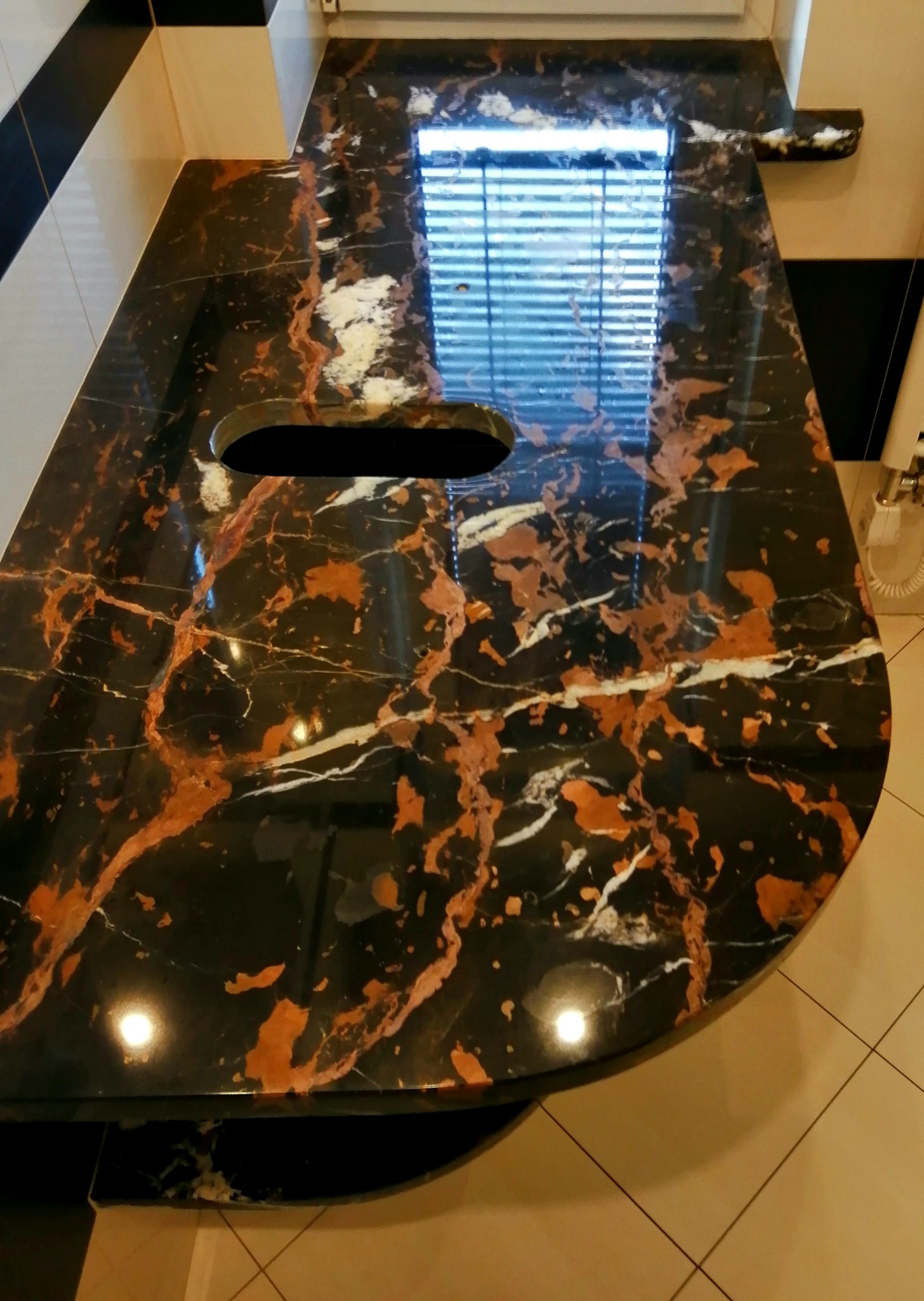Czarny blat z brązowo-białym żyłkowaniem, z otworem na zlew, odbijający światło okna w polerowanej powierzchni.