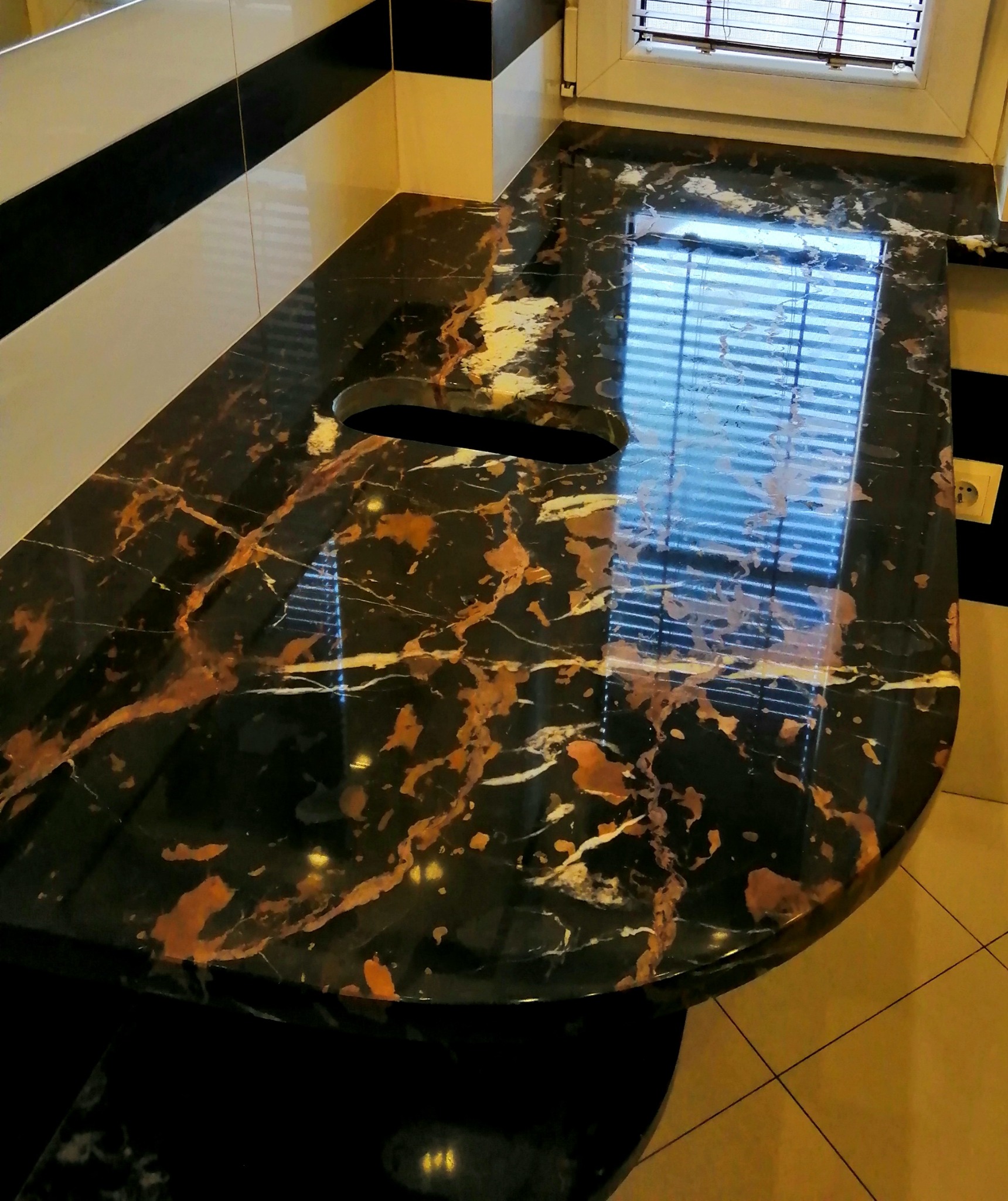 Czarny blat kamienny z brązowymi i białymi żyłami, z otworem na zlew, odbijający światło z okna w łazience z czarno-białymi płytkami.