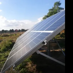 Panel słoneczny na metalowej konstrukcji, widok z dołu, na tle wiejskiego krajobrazu i błękitnego nieba z chmurami.