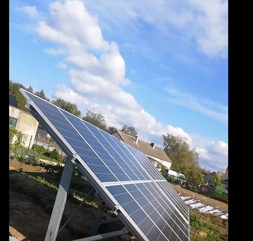 Panel słoneczny ustawiony pod kątem na metalowej konstrukcji, z widocznym budynkiem mieszkalnym w tle i błękitnym niebem z chmurami.