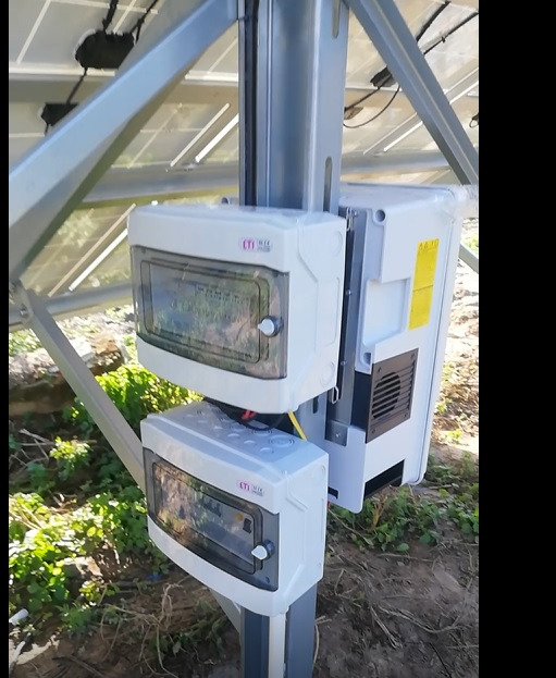 Dwa białe rozdzielacze elektryczne ETI zamontowane na metalowym słupku konstrukcji wsporczej paneli słonecznych, z widocznym inwerterem i fragmentami roślinności w tle.