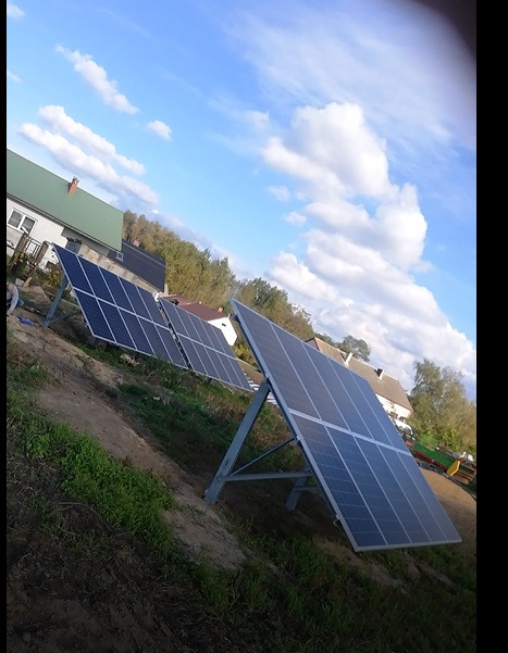 Instalacja paneli fotowoltaicznych na gruncie w wiejskiej scenerii, widoczne panele na metalowych konstrukcjach, domy w tle i błękitne niebo z chmurami.