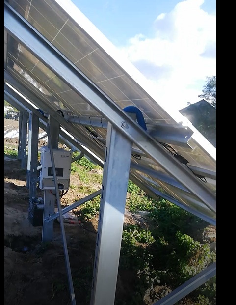 Widok od spodu na panele słoneczne zamontowane na metalowej konstrukcji, widoczny inwerter i okablowanie, tło stanowi niebo i zieleń.