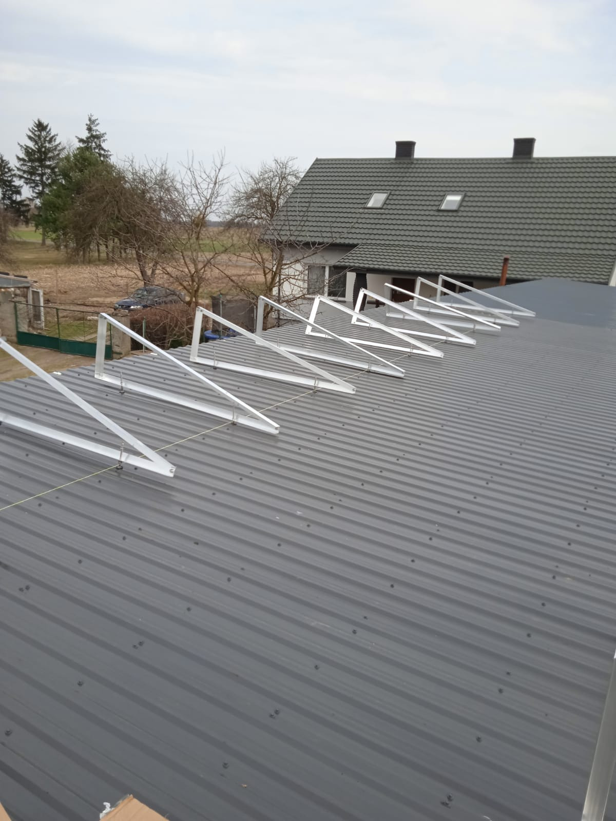 Montaż aluminiowych konstrukcji wsporczych na dachu z blachy trapezowej przygotowanych do instalacji paneli słonecznych, widok na budynek mieszkalny w tle.