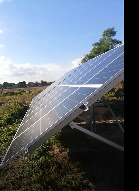 Panel słoneczny na metalowej konstrukcji, widok z dołu, na tle wiejskiego krajobrazu i błękitnego nieba z chmurami.