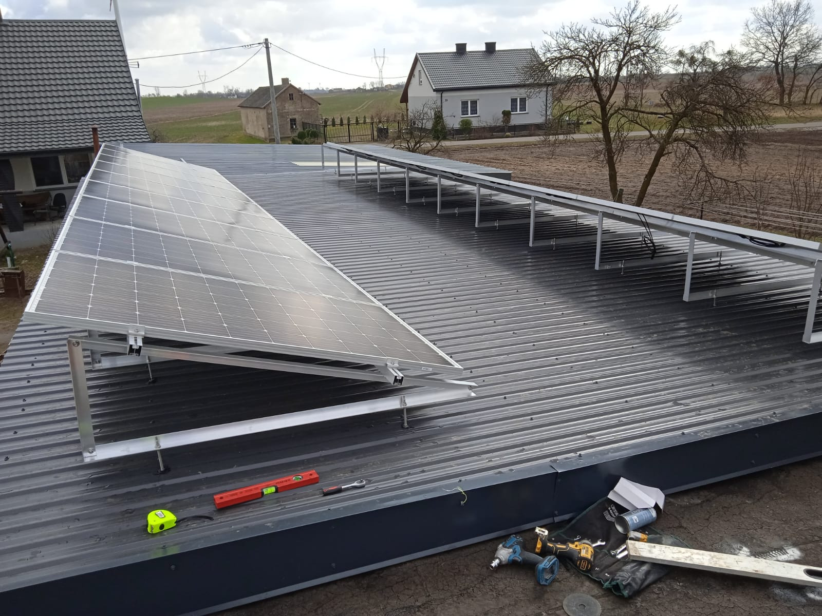 Montaż paneli słonecznych na dachu z blachy trapezowej, widoczne narzędzia pracy: poziomica, miarka, klucz, wkrętarki akumulatorowe, w tle zabudowania wiejskie pod pochmurnym niebem.