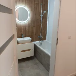 Nowoczesna łazienka z drewnopodobnymi płytkami na ścianie, okrągłym lustrem z podświetleniem LED, białą umywalką nablatową i wanną z szarym obiciem, widok zza uchylonych drzwi.