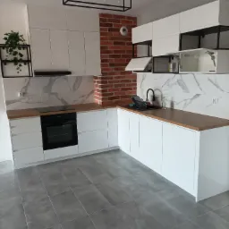 Nowoczesna kuchnia z białymi szafkami, drewnianym blatem, marmurową ścianą i ceglanym fragmentem, oświetlenie sufitowe w stylu industrialnym, szare płytki podłogowe.