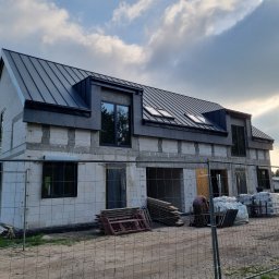 Usługi Ogólnobudowlane - Budowa domu z białych bloczków w Goleniowie, z czarnym dachem i oknami. Widoczne materiały budowlane i ogrodzenie placu budowy. Ujęcie z dołu, pochmurne niebo.