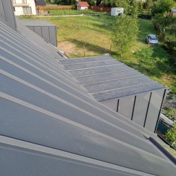 Usługi Ogólnobudowlane - Nowoczesny, szary dach z blachy na tle zielonego ogrodu i zabudowy jednorodzinnej. Widok z góry, ukazujący detale łączeń i fakturę materiału.