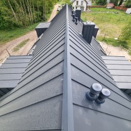 Usługi Ogólnobudowlane - Szary dach z blachy na domu jednorodzinnym, widok z góry, z kominami i wentylacją. W tle drzewa i domy. Charakterystyczne ułożenie blachy na dachu.