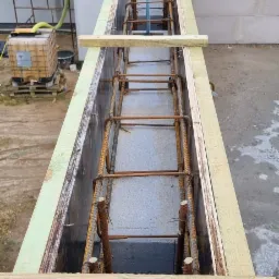 Szczegółowe ujęcie z góry szalunku zbrojeniowego wypełnionego betonem, drewniane deski stabilizujące, w tle fragment dachu z dachówki ceramicznej i pojemnik na wodę.