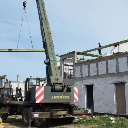 Żuraw Liebherr podnosi drewniane belki na budowę domu z betonu kom&oacute;rkowego, widoczny pracownik na g&oacute;rze konstrukcji.