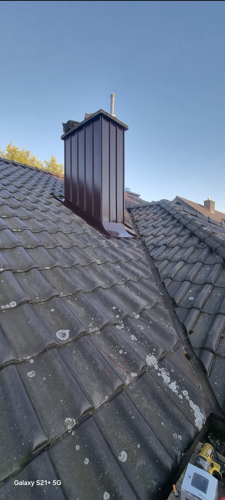 Nowy, brązowy komin z blachy na dachu pokrytym ciemną dachówką ceramiczną. Widoczne ślady zabrudzeń na dachówkach. Błękitne niebo w tle.