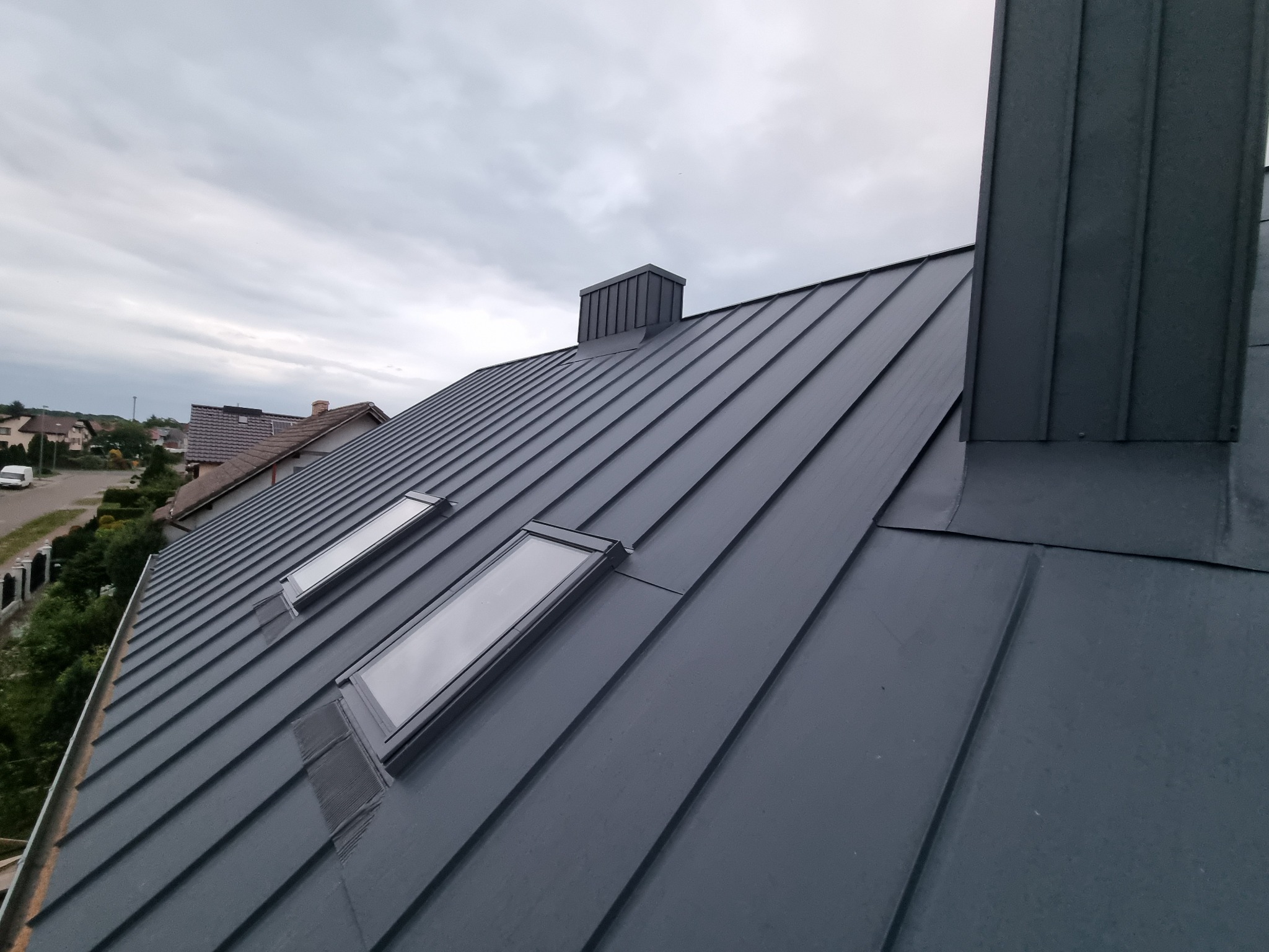 Szary dach z blachy na domu jednorodzinnym z dwoma oknami dachowymi i kominem, widok z góry, pochmurne niebo w tle.
