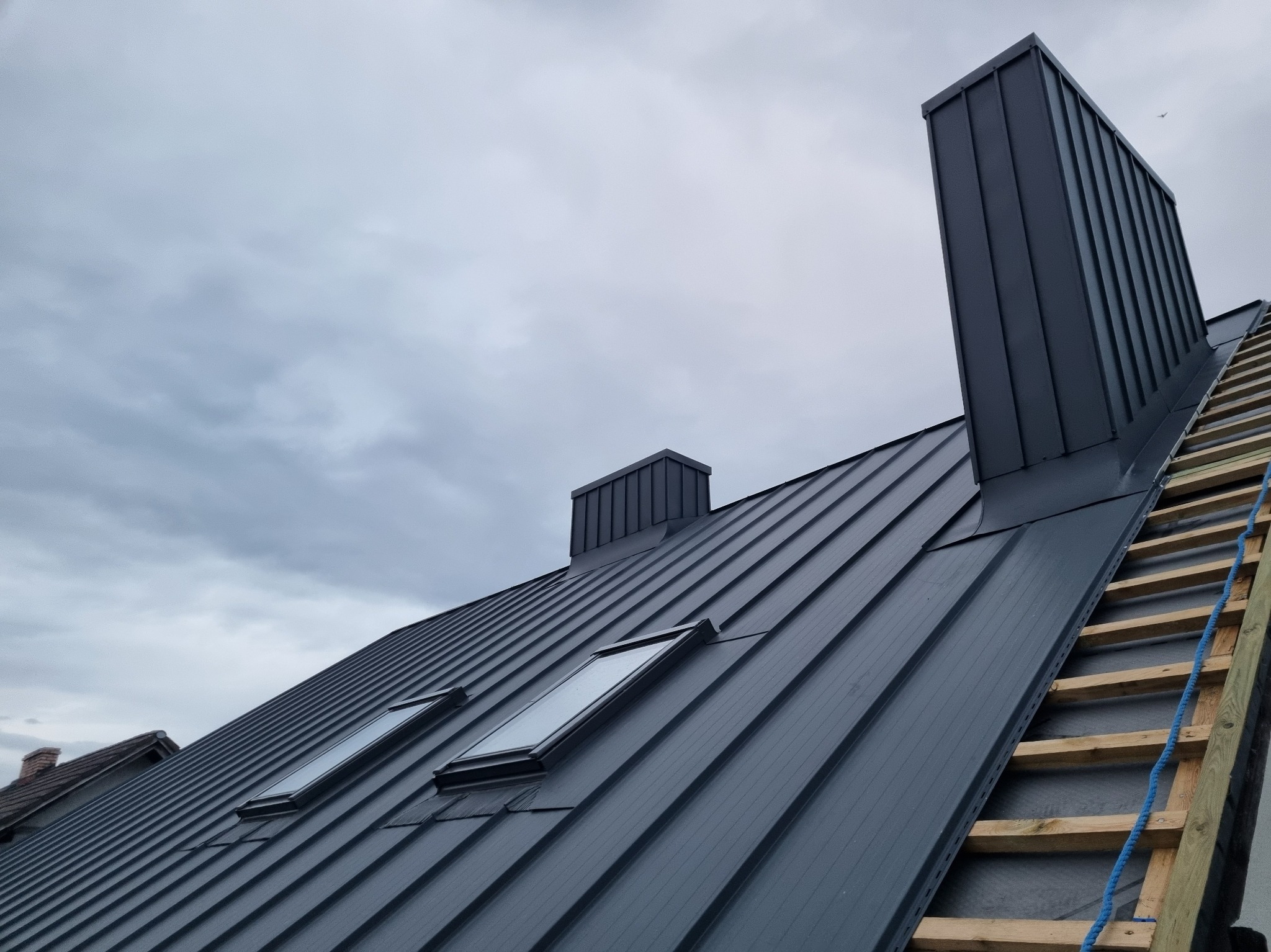 Szary dach z blachy na rąbek stojący z oknami dachowymi i kominem, widziany pod kątem. Fragment rusztowania z boku. Pochmurne niebo w tle.