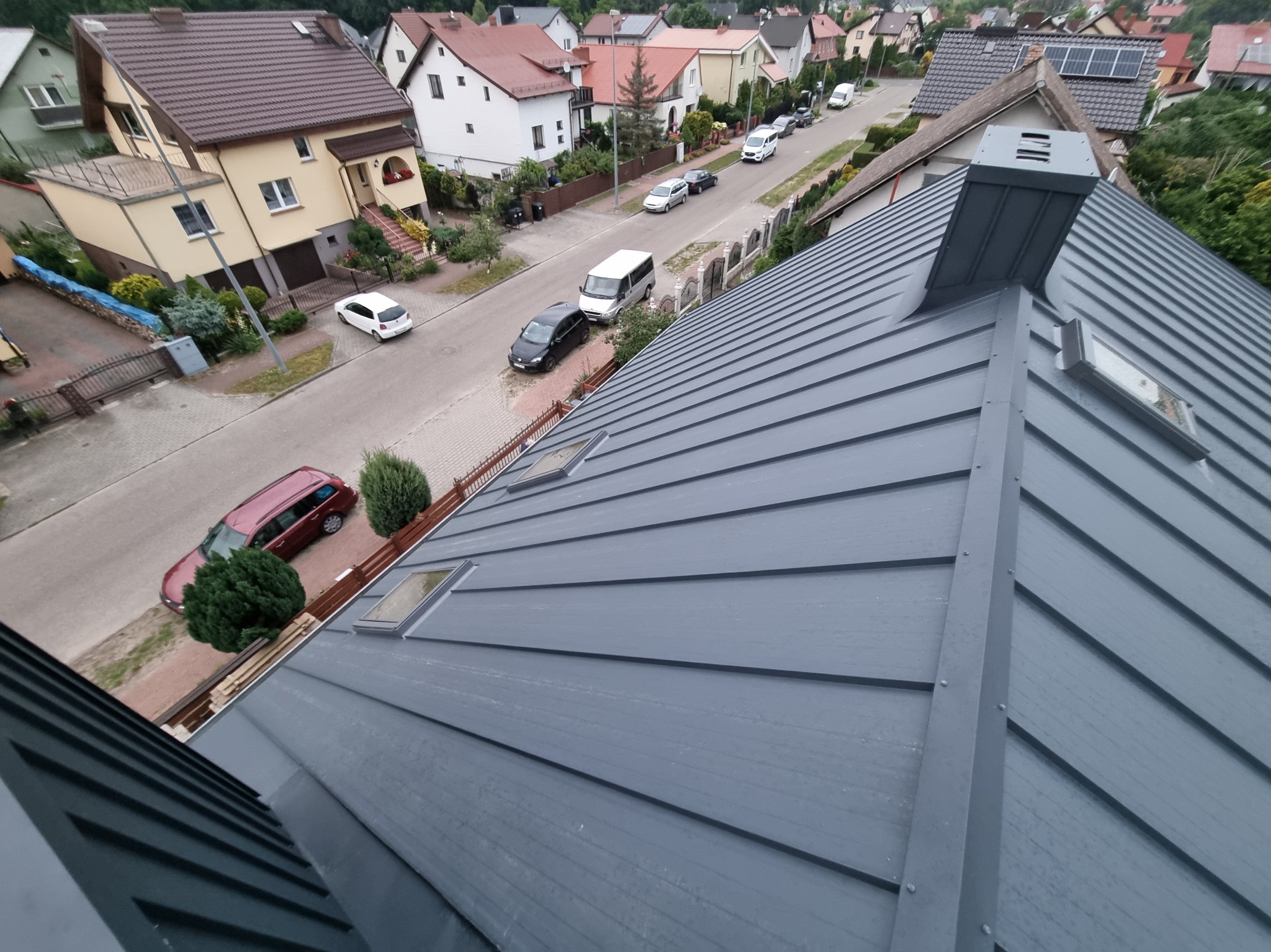 Szary, metalowy dach z oknami dachowymi i kominem, widziany z góry, z perspektywą na ulicę i okoliczne domy. Nowoczesne pokrycie dachu.