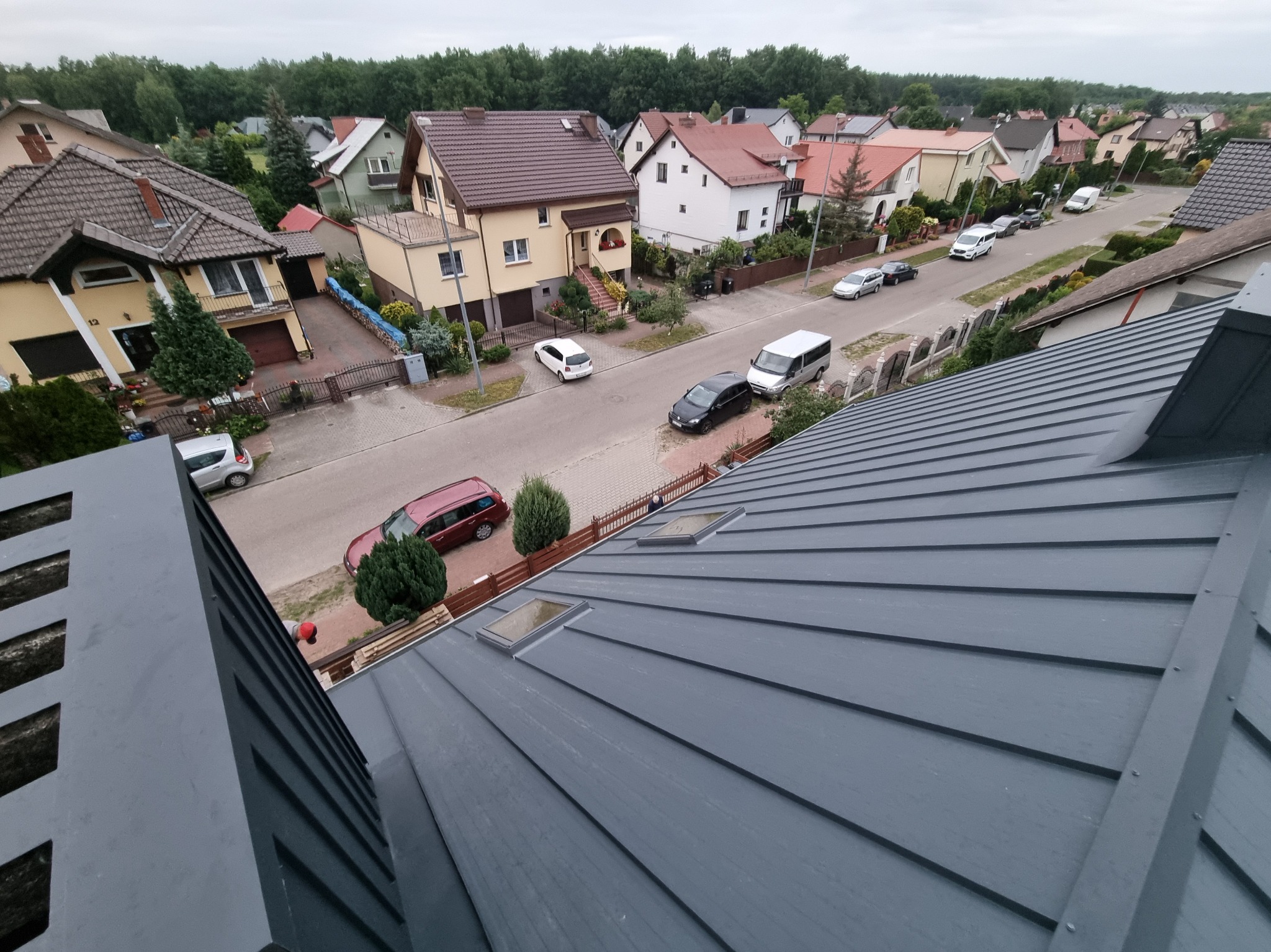 Szary, blaszany dach z oknami dachowymi, widziany z góry na tle zabudowy jednorodzinnej i drzew. Ujęcie z perspektywy dachu sąsiedniego budynku.