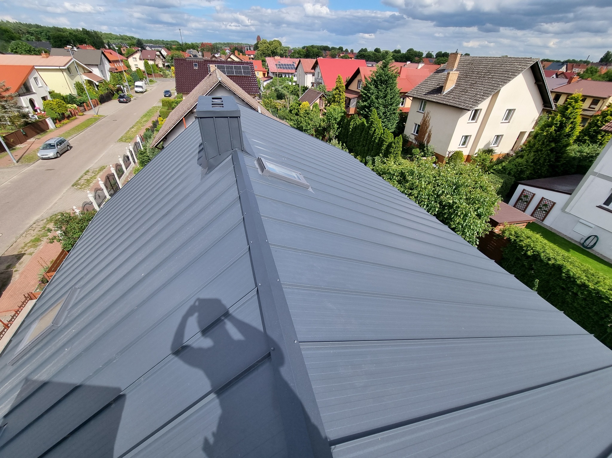 Szary dach z blachy na tle zabudowy jednorodzinnej w słoneczny dzień. Widoczny komin i okno dachowe. Cień osoby robiącej zdjęcie.