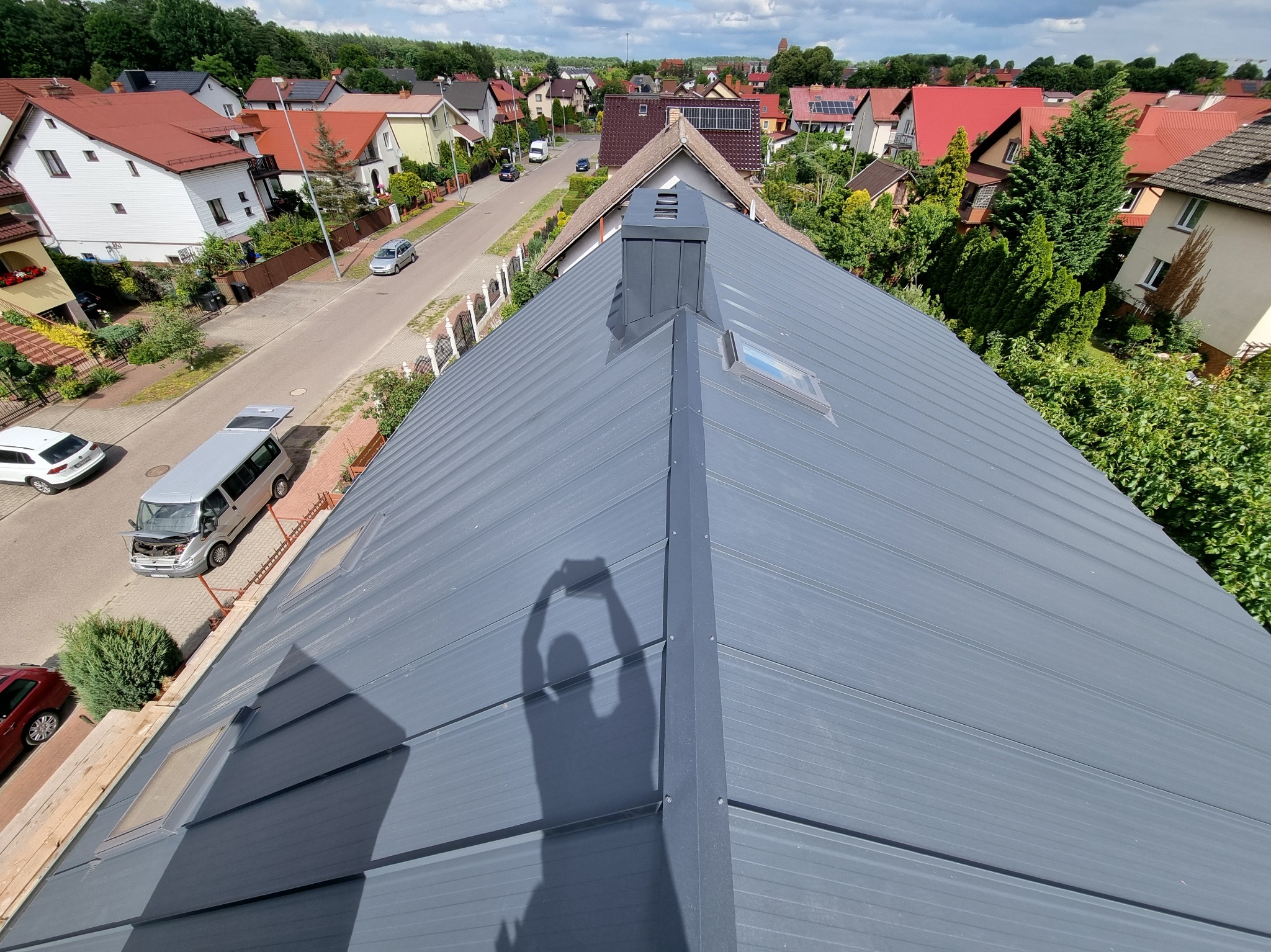 Szary dach z blachy na tle zabudowy jednorodzinnej, widok z góry z cieniem osoby robiącej zdjęcie. Widoczne okna dachowe i komin.