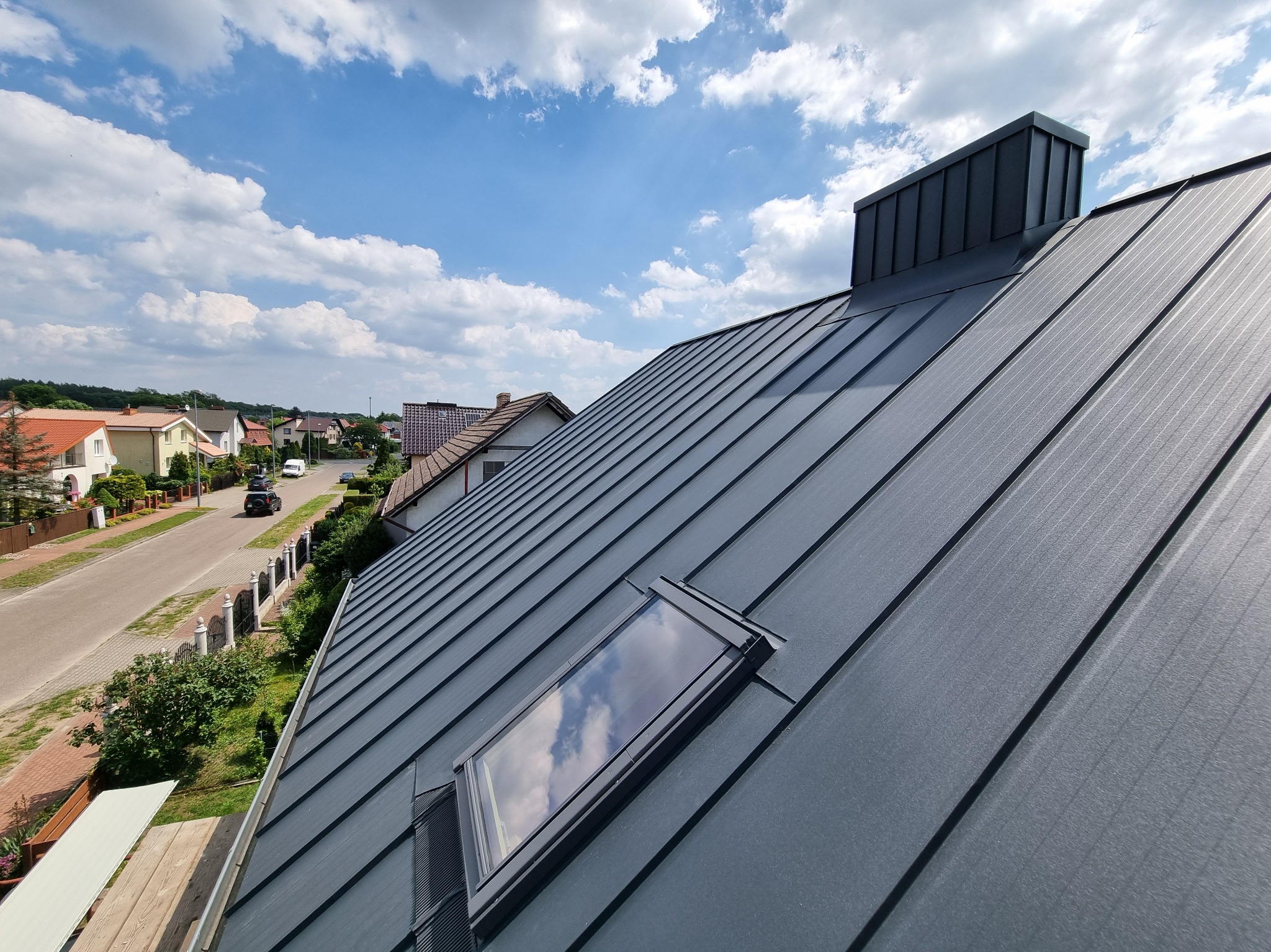Grafitowy dach z blachy na domu jednorodzinnym, widok z góry, okno dachowe odbija błękitne niebo z chmurami. W tle zabudowa mieszkalna i droga.