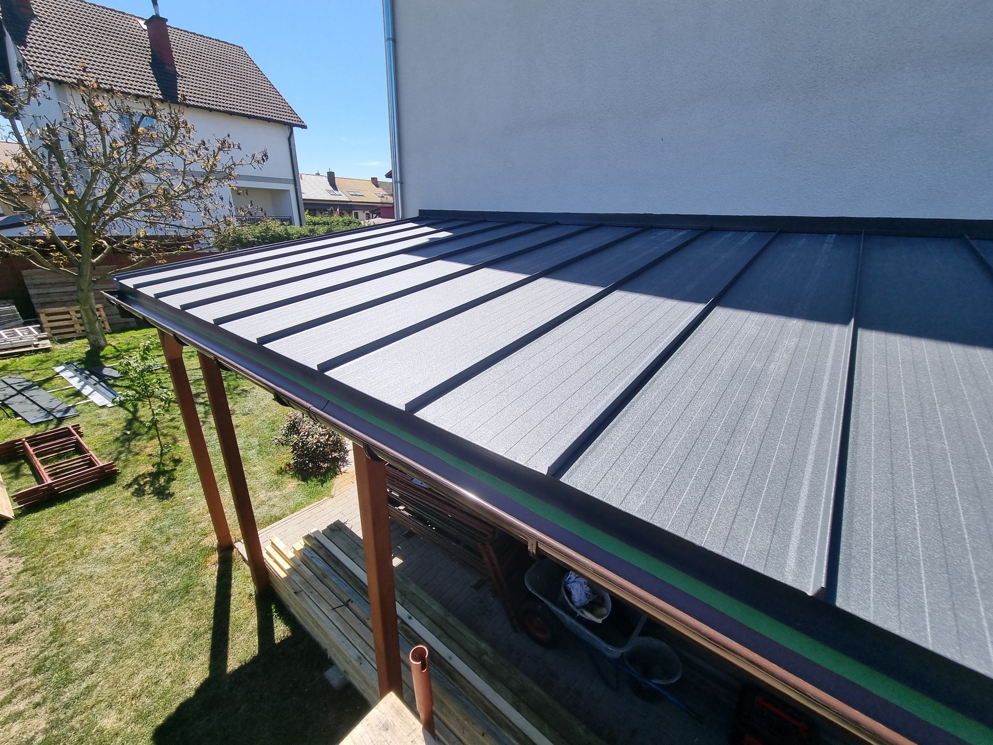 Nowoczesne, szare pokrycie dachowe z blachy trapezowej, widok z góry na fragment dachu i rynny, z elementami konstrukcji drewnianej i zieleni w tle.