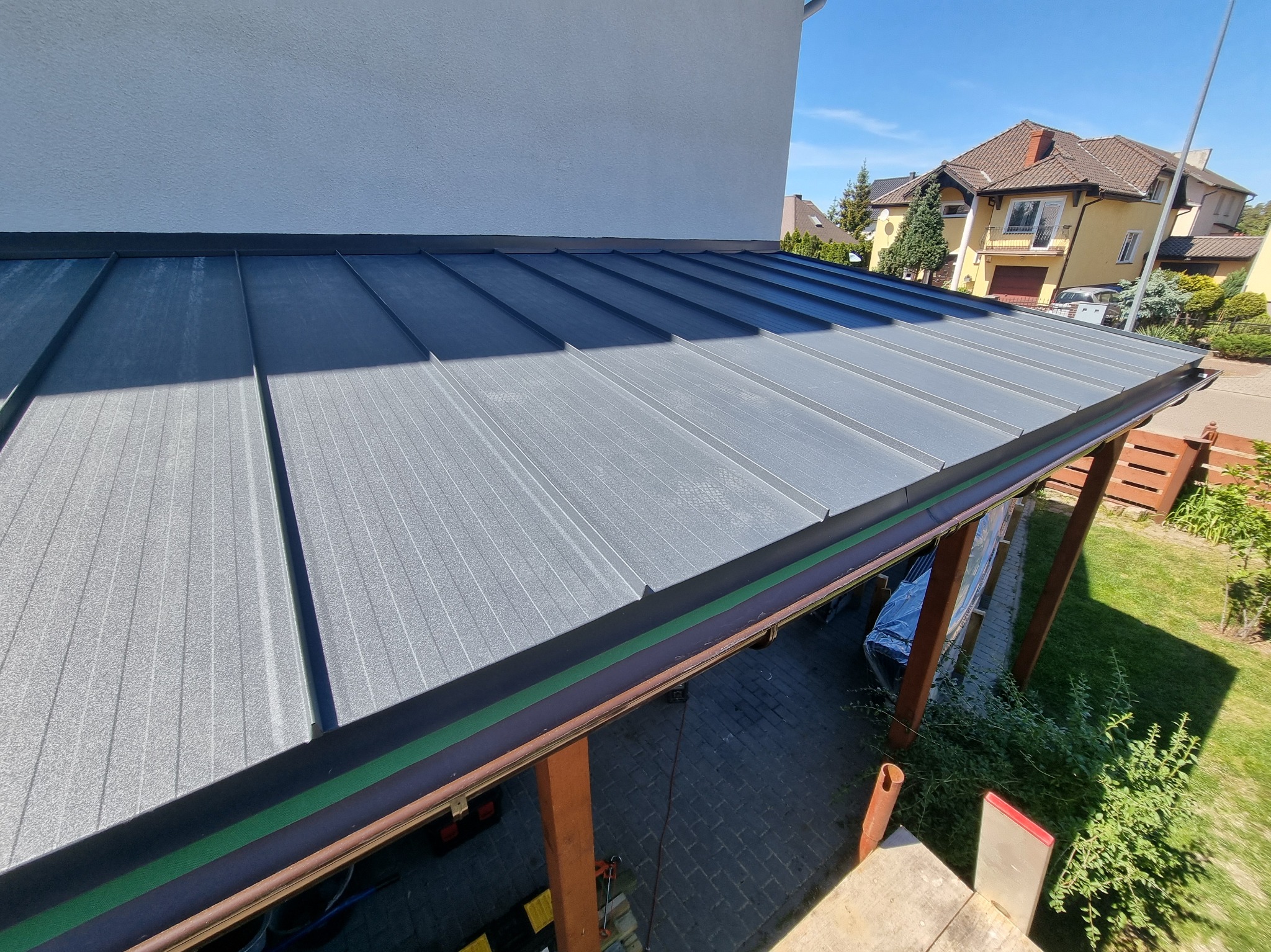 Szary dach z blachy trapezowej, widok z góry, z widocznymi elementami konstrukcyjnymi i rynną. W tle domy i błękitne niebo.