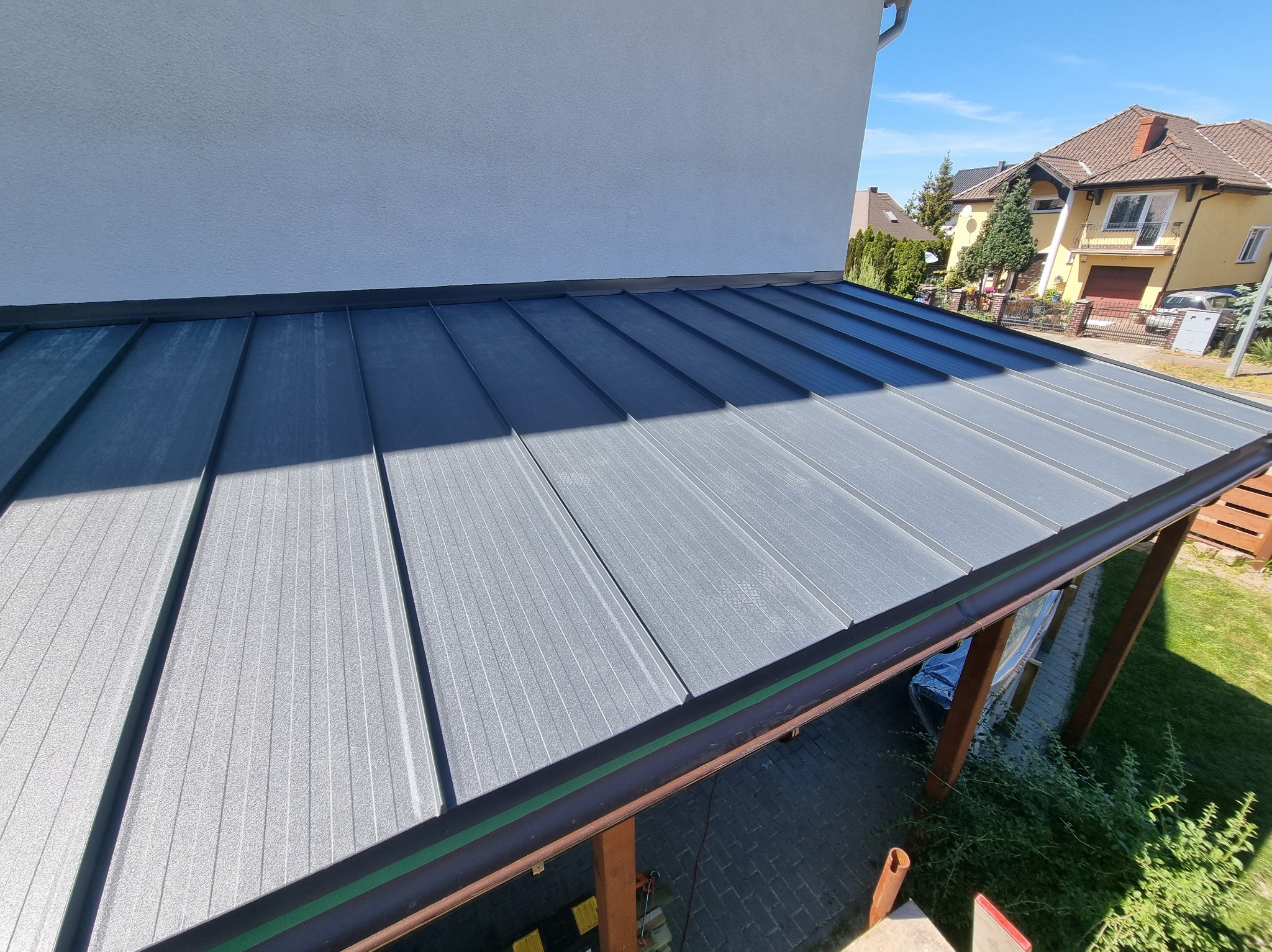 Nowoczesne, szare pokrycie dachowe z blachy na drewnianej konstrukcji w słoneczny dzień. Widok z góry, z fragmentem elewacji budynku w tle.