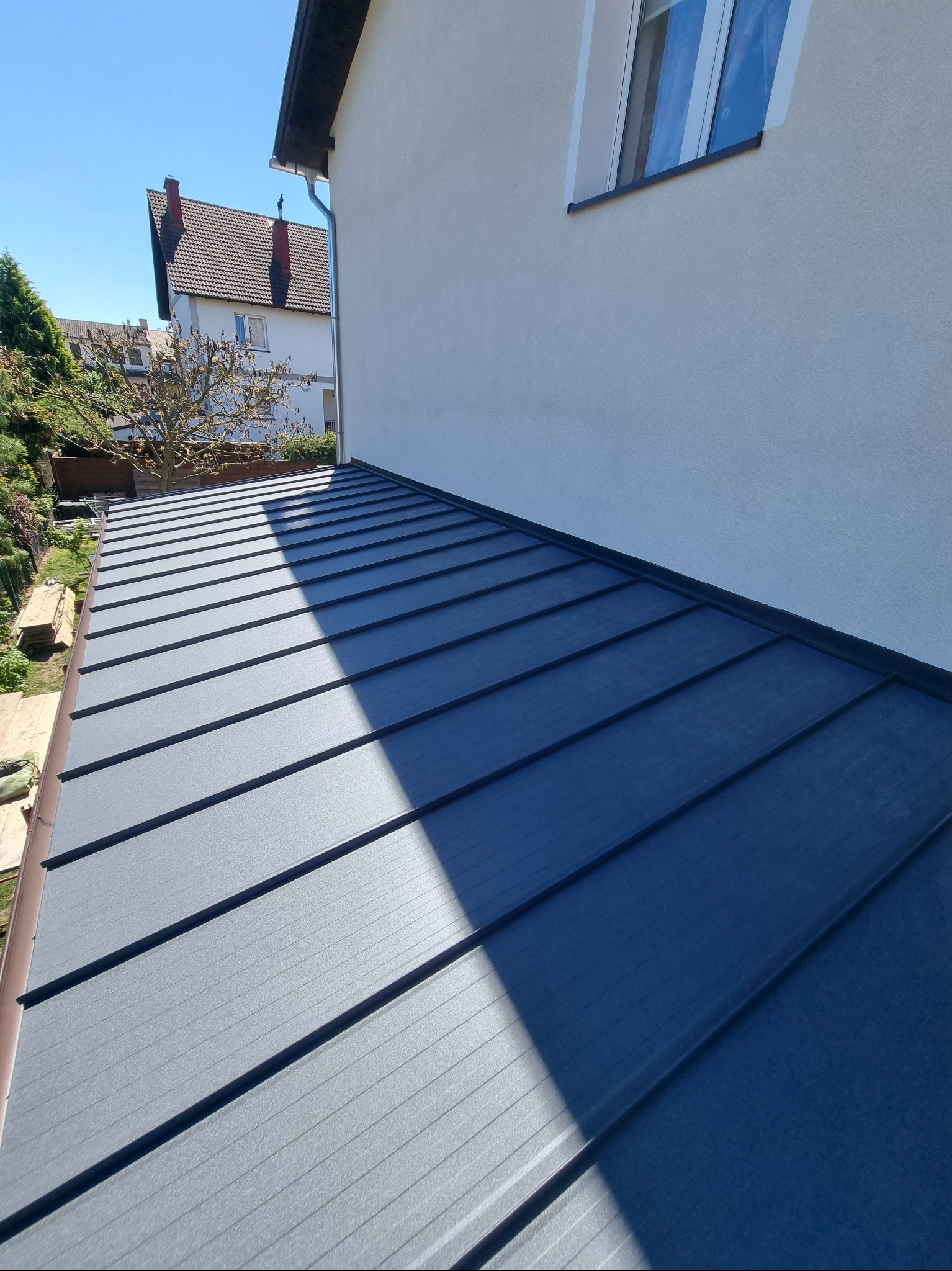 Nowoczesny, grafitowy dach z blachy trapezowej, świeżo po montażu, przylegający do jasnej ściany budynku. Widoczne brązowe rynny i fragment okna. Słoneczny dzień.