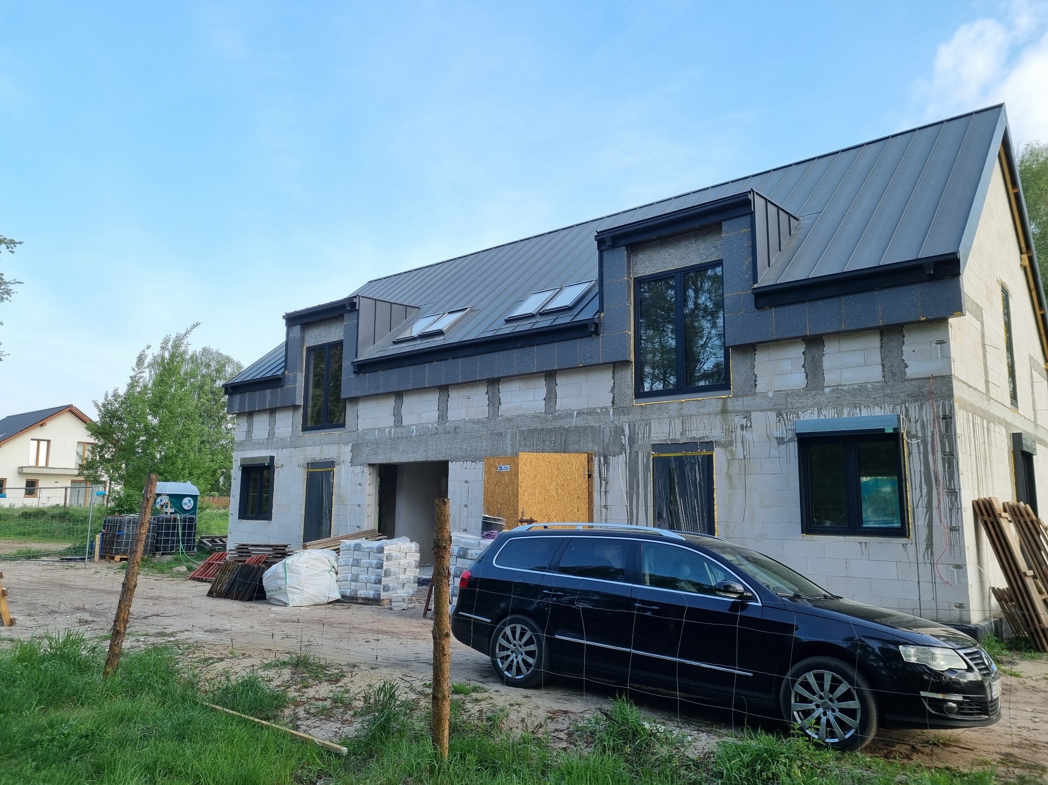 Surowy stan domu z szarego bloczka, z ciemnym dachem i oknami, otoczony materiałami budowlanymi i czarnym samochodem. Widok z zewnątrz, w trakcie budowy.