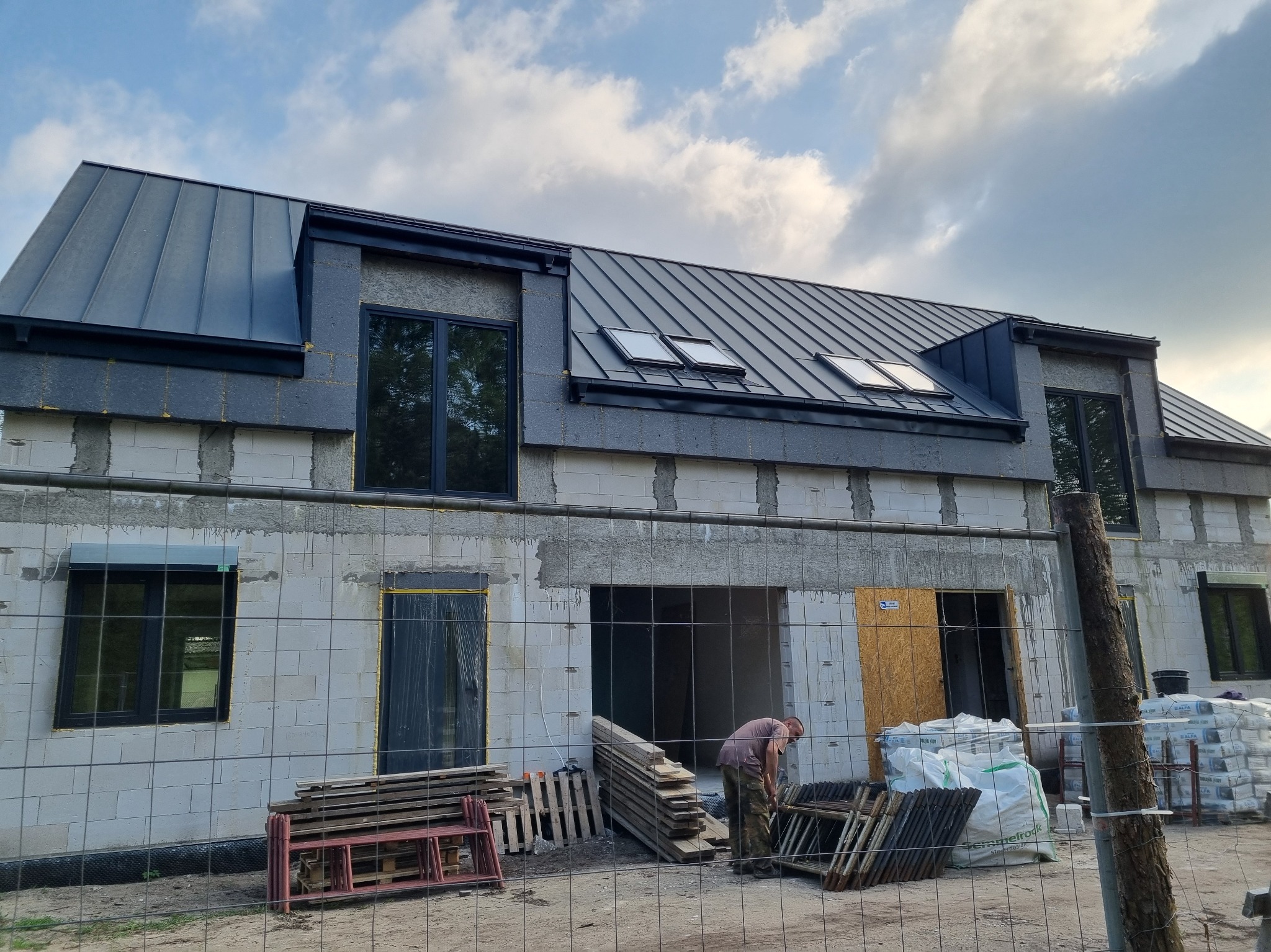 Dom w trakcie budowy z ciemnym dachem z blachy na rąbek stojący i oknami. Widoczne materiały budowlane i pracownik. Budynek z betonu komórkowego.