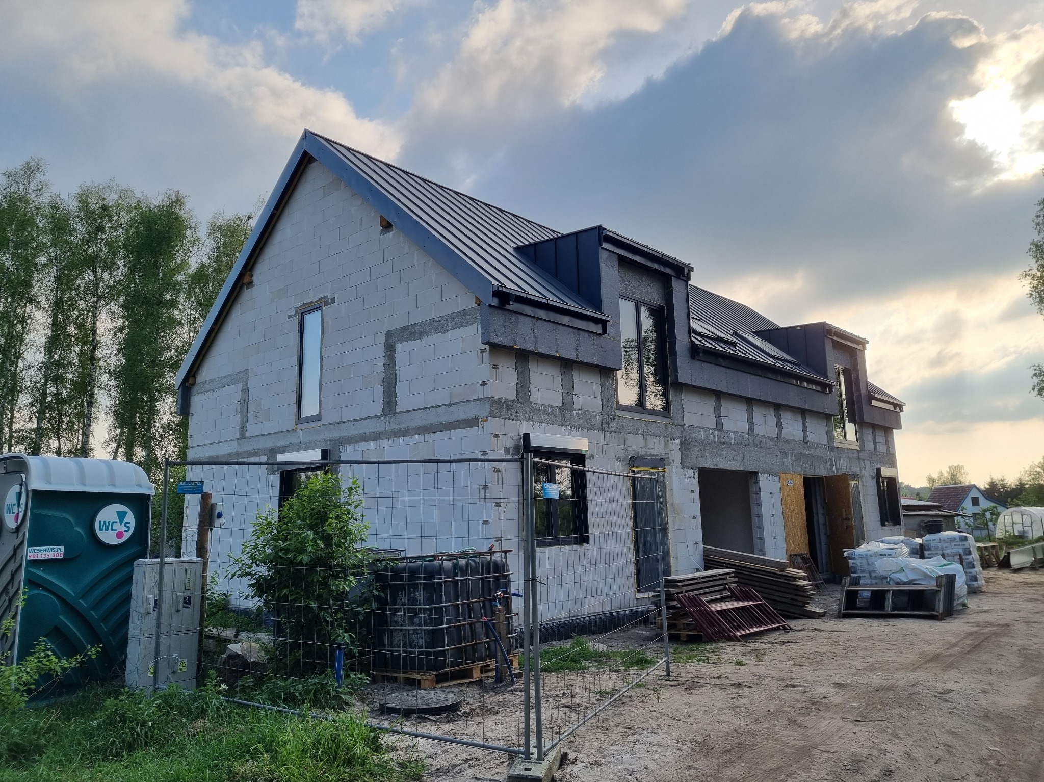 Surowy stan domu z białego pustaka z czarnym dachem, widoczne okna i elementy konstrukcyjne. Na pierwszym planie ogrodzenie, toaleta przenośna i materiały budowlane.